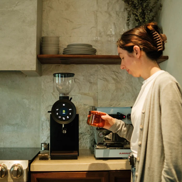 A woman in a kitchen setting holds a glass while a black coffee grinder with a digital display stands on the counter next to an espresso machine.
